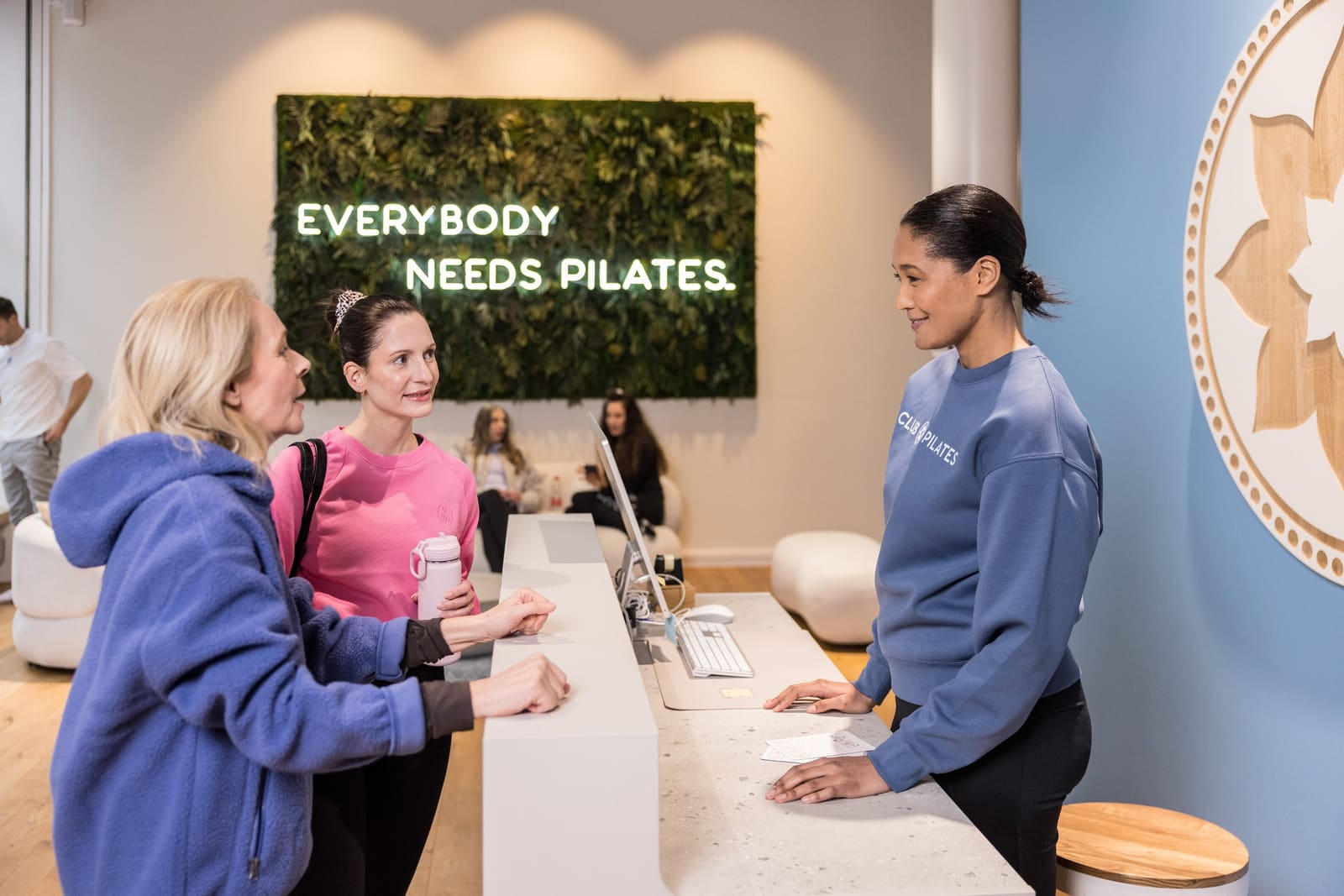 Trainerin von Club Pilates begrüßt zwei Mitglieder unterschiedlichen Alters am Empfang